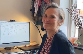 En kvinde - Anne Birgitte Gisselbæk - sidder vendt væk fra sit skrivebord og smiler mod kameraet. Bag hende ses en FRAM-model på computerskærmen.