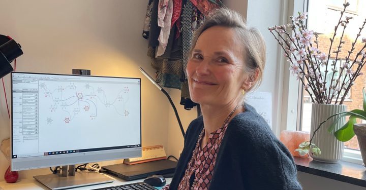 En kvinde - Anne Birgitte Gisselbæk - sidder vendt væk fra sit skrivebord og smiler mod kameraet. Bag hende ses en FRAM-model på computerskærmen.