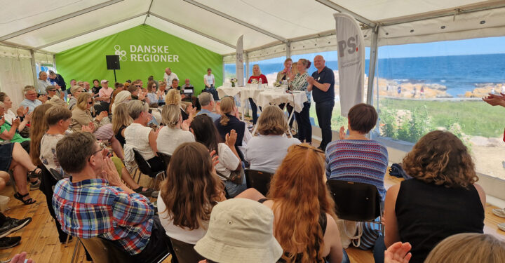 Et fyldt telt med ca. 100 tilskuere. De kigger op mod paneldeltagerne, og i baggrunden ses havet.