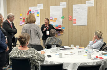 Foto fra workshop. Der står en gruppe omkring en væg, hvorpå der hænger papir og post-its.
