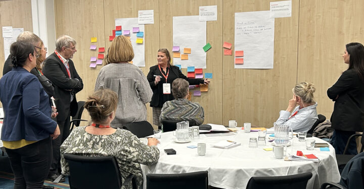 Foto fra workshop. Der står en gruppe omkring en væg, hvorpå der hænger papir og post-its.
