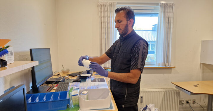 En mand kigger på et pilleglas. Han står alene i et medicinrum og foran er pilleæsker og en kasse med medicin, som han skal dispensere.