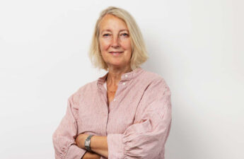 Portrætfoto af Inge Kristensen. Hun står i stribet skjorte, med armene over kors og med et smil på læben.