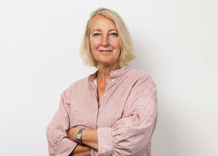 Portrætfoto af Inge Kristensen. Hun står i stribet skjorte, med armene over kors og med et smil på læben.