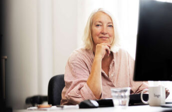 PS!-direktør Inge Kristensen sidder ved et skrivebord med computer. Hun har øjenkontakt med kameraet og smiler.