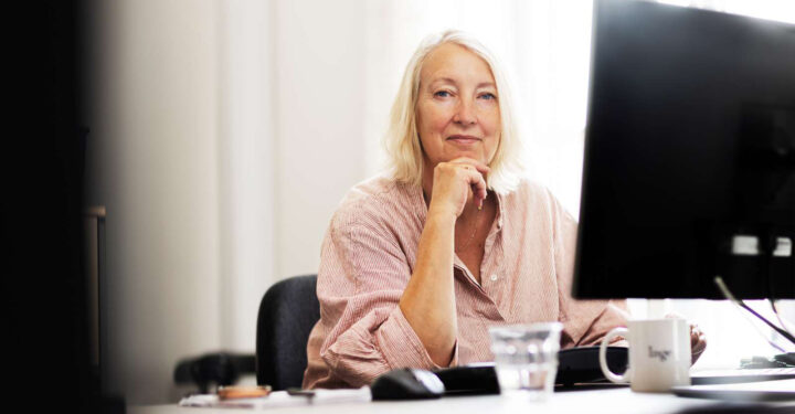 PS!-direktør Inge Kristensen sidder ved et skrivebord med computer. Hun har øjenkontakt med kameraet og smiler.