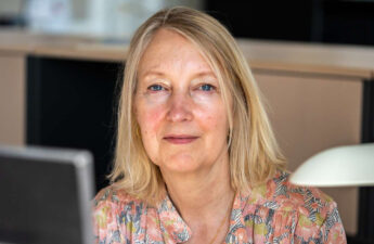 Portræt af Inge Kristensen, der sidder ved en computerskærm, men er vendt mod kameraet.