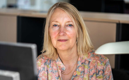 Portræt af Inge Kristensen, der sidder ved en computerskærm, men er vendt mod kameraet.