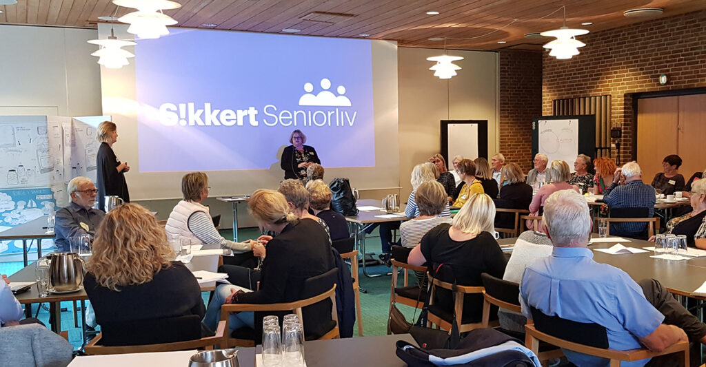 Billede af et stort mødelokale. Der sidder mennesker, både unge og ældre, ved bore med papirer. De ser mod en præsentation, hvor der står Sikkert Seniorliv.