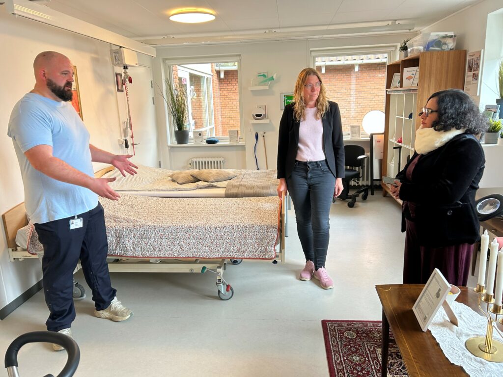 Fysioterapeut Morten fortæller om nogle af de mange hjælpemidler man kan se i Københavns Kommunes læringslejlighed på Amager. I midten Eva Steffen Hansen, leder af den kommunale hjemmepleje Amager, og til højre Dr. Kavita Kachroo.